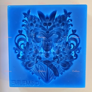 May include: Blue silicone mould with a detailed design of a woman's face with a mask and floral accents. The mould is labelled "EMLIEMSO".