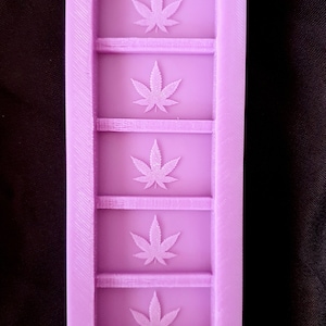Può includere: Stampo in silicone viola con cinque cavità rettangolari, ciascuna con un design a foglia di cannabis in rilievo.