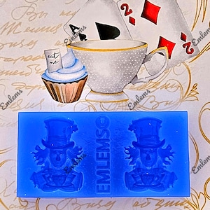 Op de afbeelding: Een blauwe siliconen mal met twee ontwerpen van de Mad Hatter. De mal is rechthoekig en heeft de tekst "EMLEMSO" erop gedrukt. De achtergrond bevat speelkaarten, een theekopje en een cupcake met de tekst "eat me".