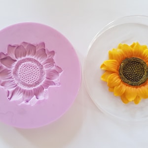 Op de afbeelding: Een siliconen mal voor het maken van zonnebloemvormige decoraties. De mal is roze en heeft een zonnebloemontwerp. Een gele zonnebloemdecoratie wordt naast de mal getoond.