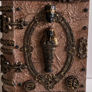 May include: An Egyptian-themed decorative box. The brown box has a textured surface and bronze-coloured metal accents. A central oval frame features a pharaoh sarcophagus and a bust. The box has a rope-like spine and metal clasps.