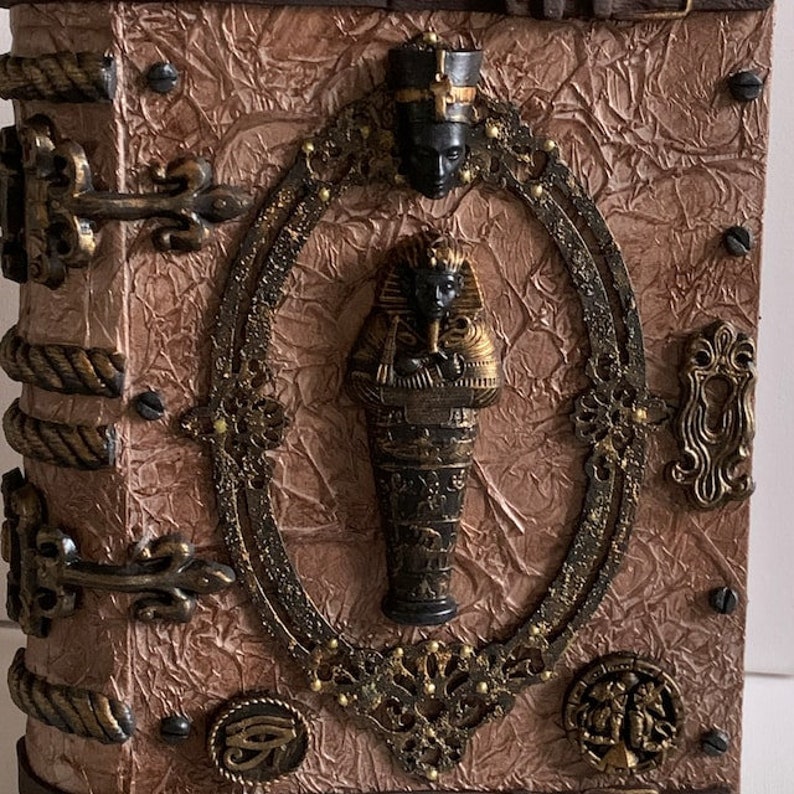 May include: An Egyptian-themed decorative box. The brown box has a textured surface and bronze-coloured metal accents. A central oval frame features a pharaoh sarcophagus and a bust. The box has a rope-like spine and metal clasps.