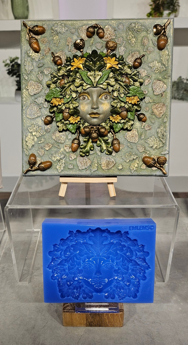 May include: A blue silicone mold for making a green woman's face with leaves and acorns. The mold is on a wooden stand and the text "EMILEMSCO" is visible on the mold.