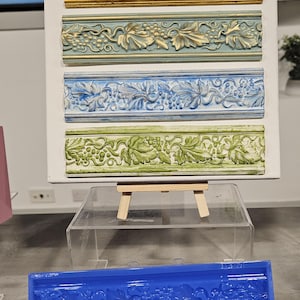 May include: A blue silicone mould for creating decorative trim with a grapevine design. The mould is shown on a clear acrylic surface. The mould is 25.4 cm long and 5.1 cm wide.