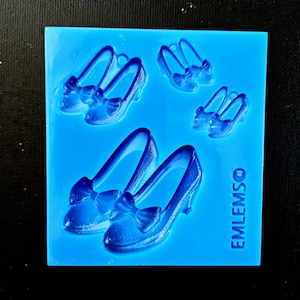 May include: A blue silicone mold featuring multiple pairs of high-heeled shoes with bow accents. The shoes are arranged in different sizes and orientations. The text "EMLEMSO" is printed on the side of the mold.