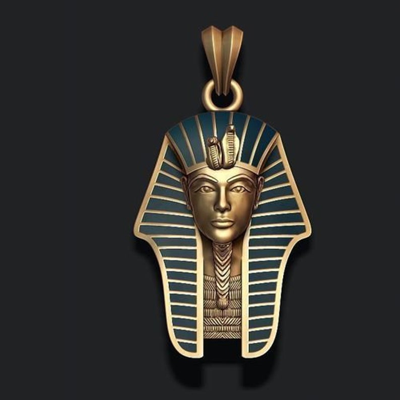May include: A gold-coloured pendant depicting the bust of an Egyptian pharaoh, with a blue and gold headdress. The pendant has a detailed design and a loop for a chain. The background is a dark grey.