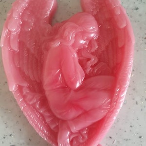 May include: A pink angel figurine with wings outstretched, the angel is kneeling with its head bowed. The figurine is made of a smooth, shiny material.