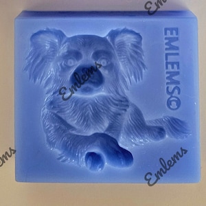 May include: A light blue, rectangular mold featuring a detailed relief of a koala bear. The koala is depicted in a seated position. The text "EMLEMSO" is visible on the right side of the mold. The word "Emlems" is also visible.