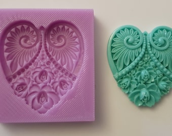 May include: A purple silicone mould shaped like a heart with a detailed floral design. The mould is used to create a teal heart-shaped pendant with a similar floral design.