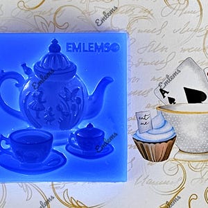 Puede incluir: Molde de silicona azul para hacer una tetera, una taza de té y un platillo. El molde está sobre una superficie blanca con un fondo de escritura cursiva.