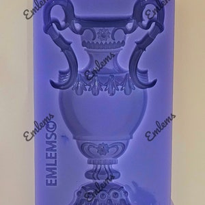 May include: Purple silicone mould for making a decorative vase with ornate details. The mould is rectangular and has the word "Emblems" printed on all sides.