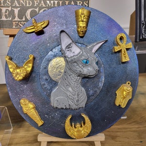 May include: A circular decorative artwork with a grey Sphynx cat at its centre, surrounded by gold Egyptian symbols. The background is dark blue with a starry effect. It is displayed on a small wooden easel.