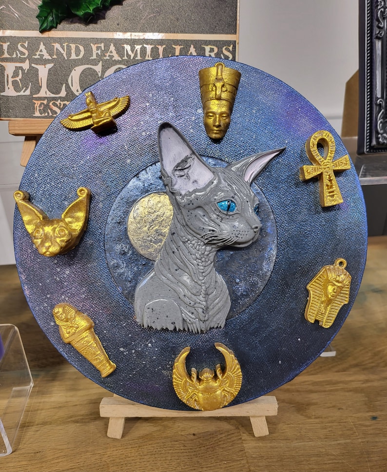 May include: A circular decorative artwork with a grey Sphynx cat at its centre, surrounded by gold Egyptian symbols. The background is dark blue with a starry effect. It is displayed on a small wooden easel.