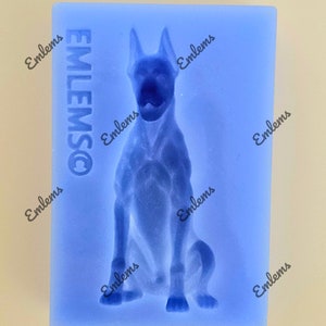 May include: A blue silicone mould depicting a seated dog with an open mouth. The mould has the word "EMLEMSCO" printed vertically on the left side. The dog's form is detailed, with visible ears and legs. The background is a light beige colour.