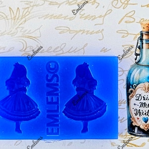 Op de afbeelding: Een blauwe siliconen mal met twee indrukken van een meisje in een blauwe jurk. De mal is gelabeld "EMLEMSO". Een blauwe glazen fles met een kurkstop en een etiket met de tekst "Drink Me Aivided" wordt naast de mal getoond.