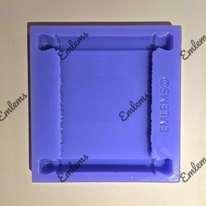 May include: Purple silicone mould for making a scroll-shaped object. The mould has a rectangular shape with rounded corners and a raised design around the edges. The mould is labelled "EMLEMSO" in the bottom right corner.