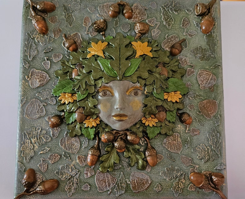 May include: A 3D mixed media artwork featuring a woman's face surrounded by green leaves, acorns, and yellow flowers. The artwork is painted on a canvas with a textured background.