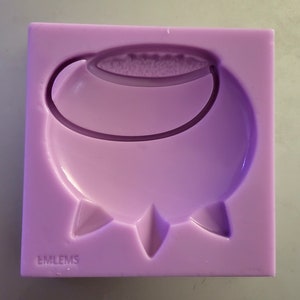 Könnte beinhalten: Eine lavendelfarbene Silikonform in quadratischer Form mit einem Kesseldesign. Der Kessel hat einen abgerundeten Körper, einen gebogenen Griff und drei spitze Füße. Der Text "EMLEMS" ist in der linken unteren Ecke aufgedruckt.