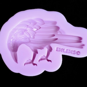 May include: Purple silicone mould for making a detailed raven or crow figurine. The mould has a smooth surface and the raven is facing left with its wings spread. The mould is marked with the word "EMLENSO".