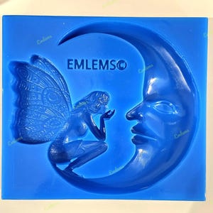 May include: A blue silicone mould depicting a fairy with detailed wings and a crescent moon with a face. The text "EMLEMSO" is visible. The fairy is kneeling and blowing towards the moon. The mould is a solid blue colour.