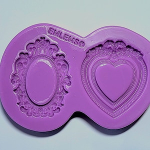 May include: Purple silicone mold with two cavities. One cavity is shaped like an oval frame with ornate details. The other cavity is shaped like a heart frame with ornate details. The mold is labeled "EMLEMSO".