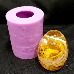 Może przedstawiać: Fioletowa silikonowa forma do robienia szklanych jajek. Forma jest cylindryczna i ma gładką, fakturowaną powierzchnię. Szklane jajko jest żółte z białymi smugami i pęcherzykami powietrza.