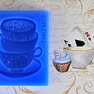 May include: A blue silicone mold of a stack of three teacups and a saucer. The mold is on a white background with a teacup, cupcake, and playing cards.
