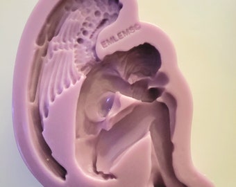 May include: Purple silicone mold of an angel with wings, sitting with its head down, holding something in its hands. The mold has the word "EMLEMOO" embossed on the side.