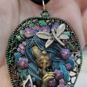 May include: A green and black metal pendant necklace featuring a woman's face with blue hair, surrounded by purple flowers and butterflies. The pendant is shaped like a leaf.