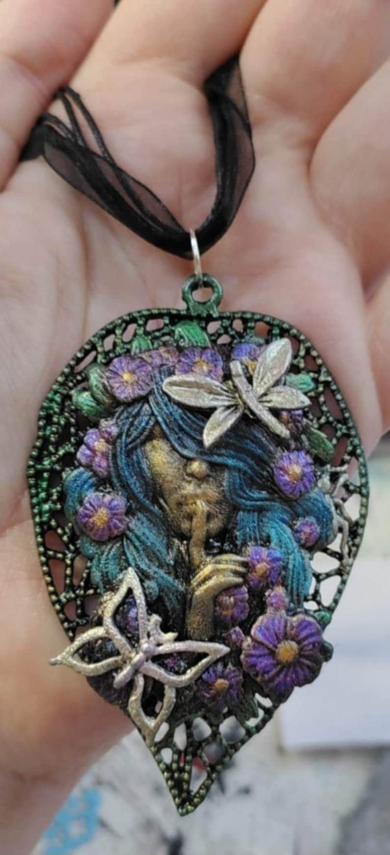 May include: A green and black metal pendant necklace featuring a woman's face with blue hair, surrounded by purple flowers and butterflies. The pendant is shaped like a leaf.