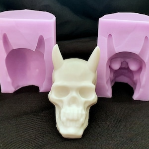 May include: A white resin skull with horns, sitting in front of two purple silicone molds. The molds are open and show the inside of the skull shape.
