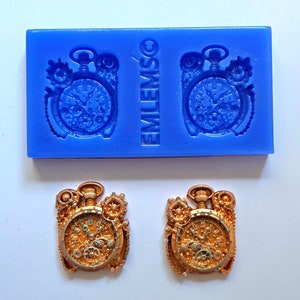 Op de afbeelding: Een blauwe siliconen mal met de tekst "EMLEM SO" en twee zakhorloge-afdrukken. Daaronder twee afgewerkte, gedetailleerde zakhorloge-hangers met goud- en zilverkleuren, met ingewikkelde tandwielen en ontwerpen.