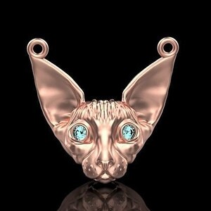 May include: A rose gold pendant shaped like a Sphynx cat's head, featuring large ears and light blue gemstone eyes. The pendant has loops at the top for a chain. The background is black.