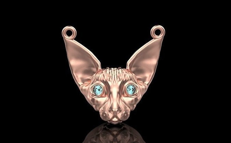 May include: A rose gold pendant shaped like a Sphynx cat's head, featuring large ears and light blue gemstone eyes. The pendant has loops at the top for a chain. The background is black.