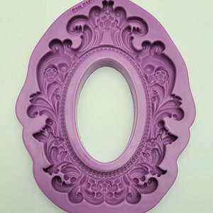 May include: Purple silicone mould shaped like an ornate oval frame with a raised design. The mould is for making decorative cakes or other edible creations.