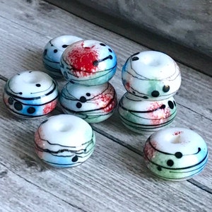 May include: A group of eight white glass beads with abstract designs in black, red, blue, and green. The beads are round and have a hole in the center.