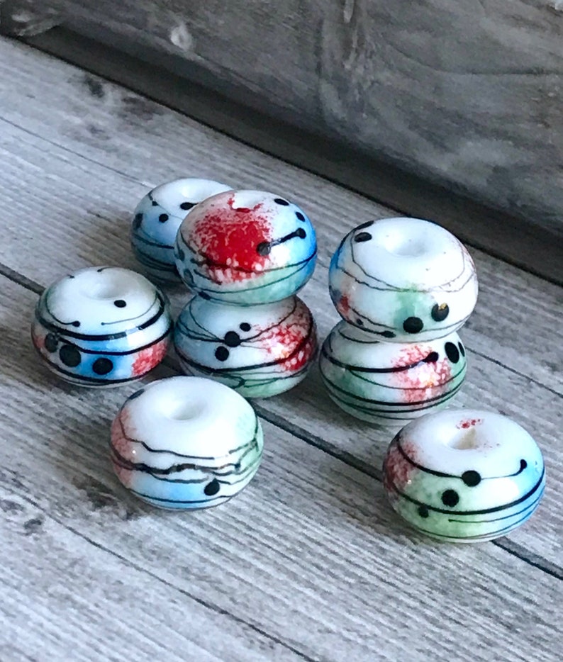 May include: A group of eight white glass beads with abstract designs in black, red, blue, and green. The beads are round and have a hole in the center.