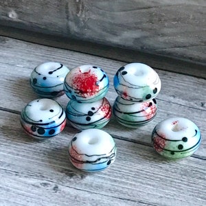 May include: A group of ten white glass beads with black, red, blue, and green abstract designs. The beads are round and have a hole in the center.