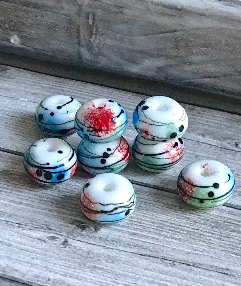 May include: A group of ten white glass beads with black, red, blue, and green abstract designs. The beads are round and have a hole in the center.