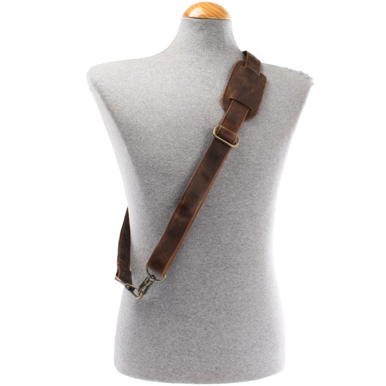 May include: A brown leather shoulder strap with a padded shoulder rest and brass-coloured metal hardware. The strap is displayed on a grey mannequin, showcasing its design and construction. The leather has a distressed appearance.