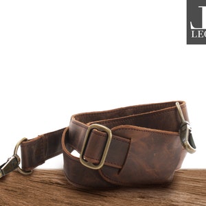 May include: A brown leather strap with brass-coloured metal hardware. The strap has a buckle, a swivel clasp, and a D-ring. The leather has a distressed appearance, and the strap appears to be for a bag or camera.