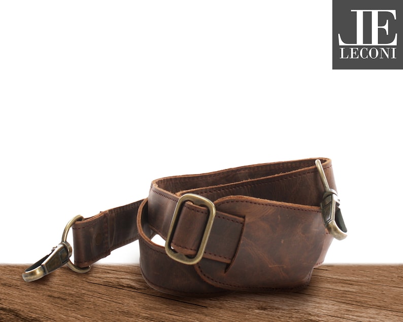 May include: A brown leather strap with brass-coloured metal hardware. The strap has a buckle, a swivel clasp, and a D-ring. The leather has a distressed appearance, and the strap appears to be for a bag or camera.