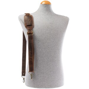 May include: A brown leather shoulder strap with a padded shoulder rest and adjustable length. The strap features brass-coloured metal hardware, including a buckle and swivel clasps for attachment. The leather has a distressed appearance.