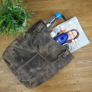 May include: A brown leather tote bag with a zippered pocket and a buckle closure. The bag is filled with various items, including a water bottle, a magazine, and a blue device.