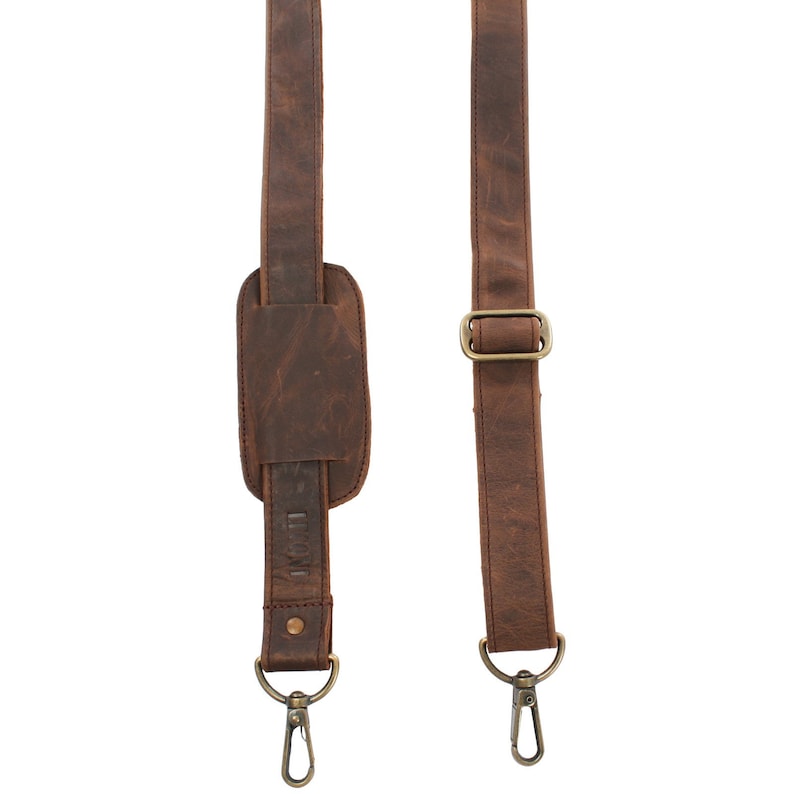 May include: Close-up of a brown leather shoulder strap with a padded shoulder rest. The strap features metal hardware, including a buckle and two swivel clasps. The leather has a distressed appearance.