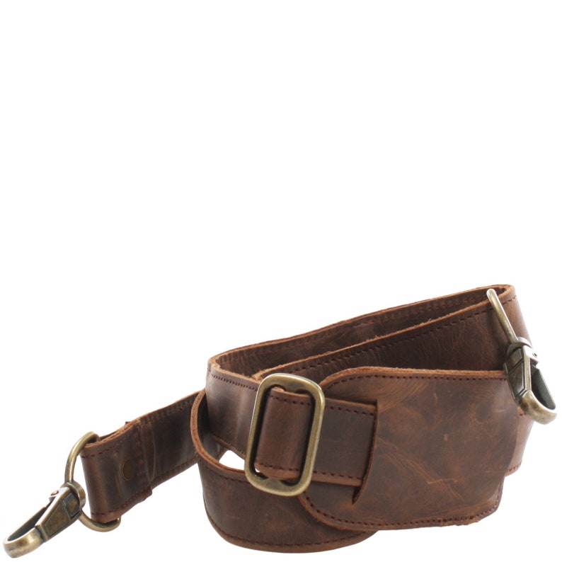 May include: A brown leather bag strap with brass-coloured hardware. The strap features a buckle, a swivel clasp, and a D-ring for attachment. The leather has a distressed appearance.