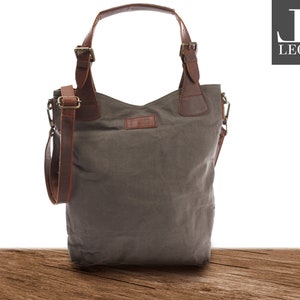 May include: A gray canvas tote bag with brown leather straps and handles. The bag has a zipper closure and a small pocket on the front. The bag is sitting on a wooden surface.