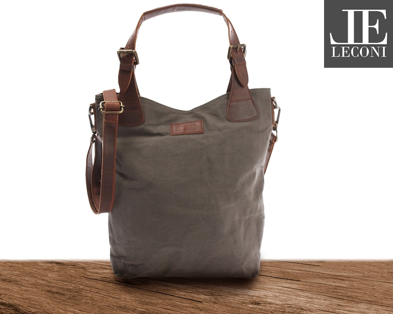 May include: A gray canvas tote bag with brown leather straps and handles. The bag has a zipper closure and a small pocket on the front. The bag is sitting on a wooden surface.