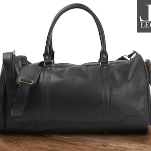 Pode incluir: Bolsa de viagem de couro preto com uma alça de ombro e um bolso com zíper lateral. A bolsa tem uma alça superior e uma etiqueta da marca que diz "JE Leconi".