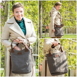 May include: A gray canvas tote bag with brown leather accents and a long shoulder strap. The bag is shown being held by a person wearing a beige coat.